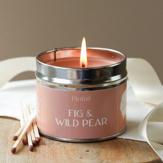Candle in a tin labeled 'Fig & Wild Pear' with matches on a wooden surface.