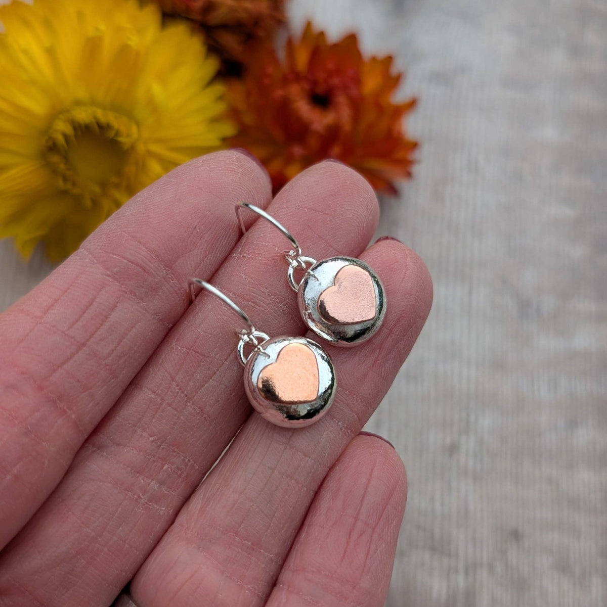 A pair of small heart-shaped dangle earrings held on a person's fingers, with yellow and orange flowers blurred in the background.
