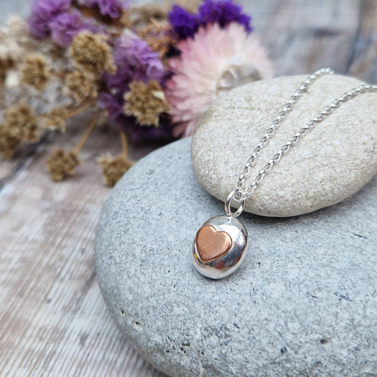 A silver chain necklace with a small heart-shaped pendant, one half silver and the other half rose gold, resting on a smooth gray stone with dried flowers in the background.