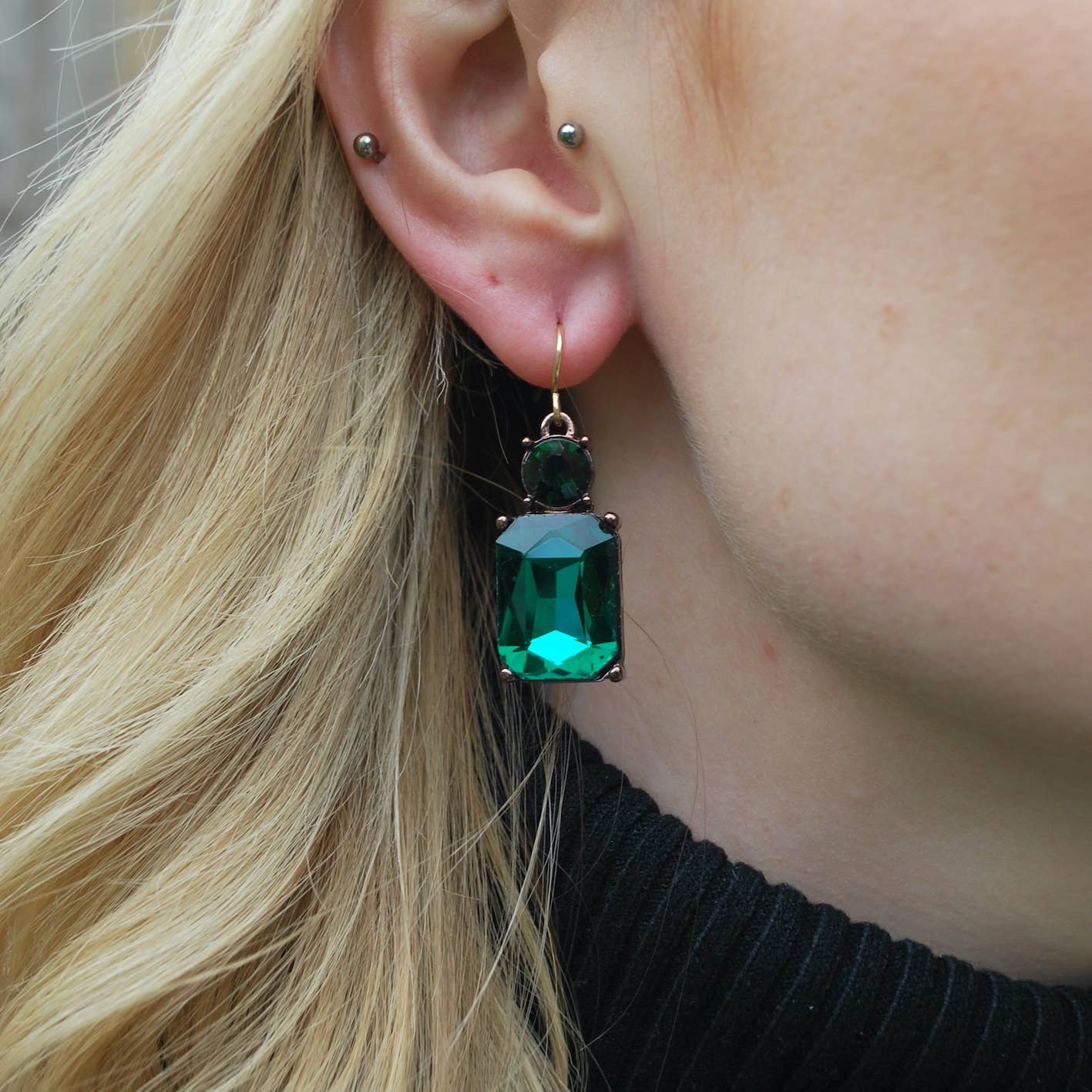 Close-up of an ear wearing a green gemstone earring with blonde hair and black top.