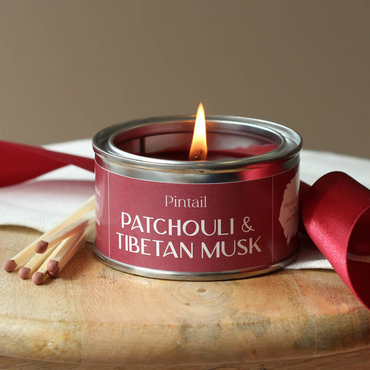 Candle in a red tin labeled 'Patchouli & Tibetan Musk' with matches and a ribbon on a wooden surface.