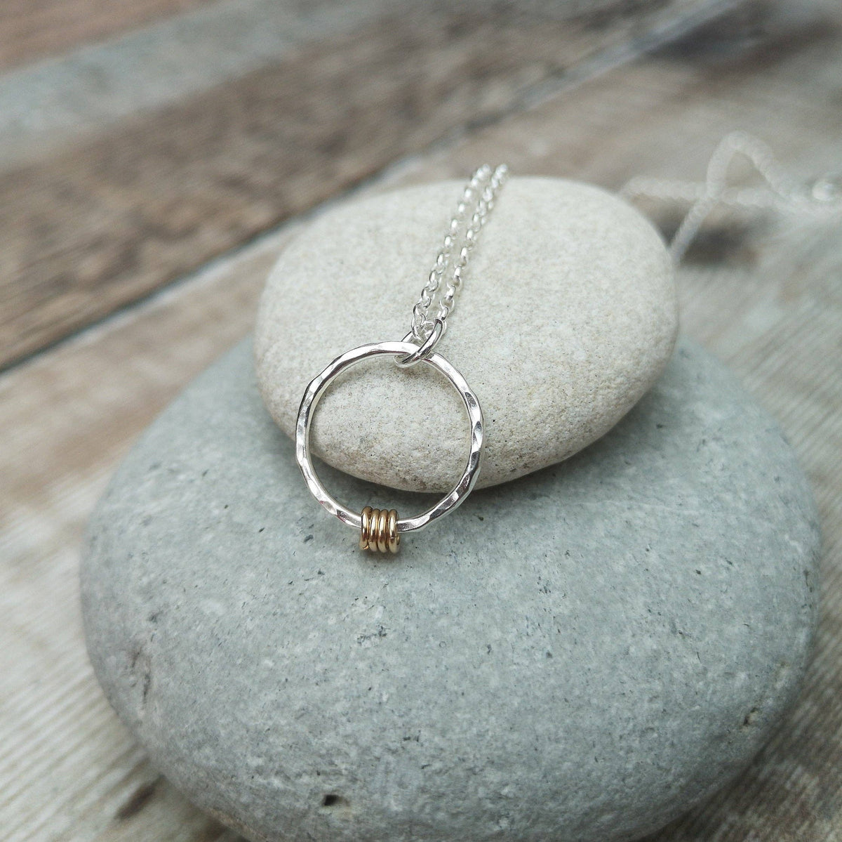 A silver ring pendant necklace with a small gold accent bead, resting on a smooth gray stone over a rustic wooden surface.