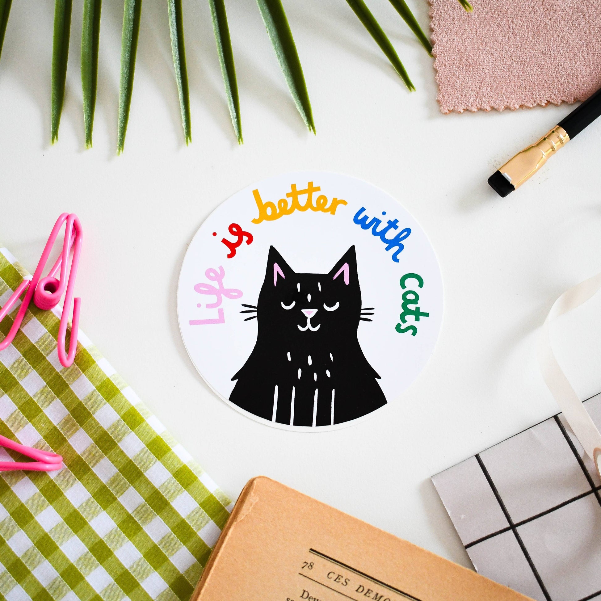 Circular sticker with a black cat illustration and 'Life is better with cats' text on a white surface with stationery items.
