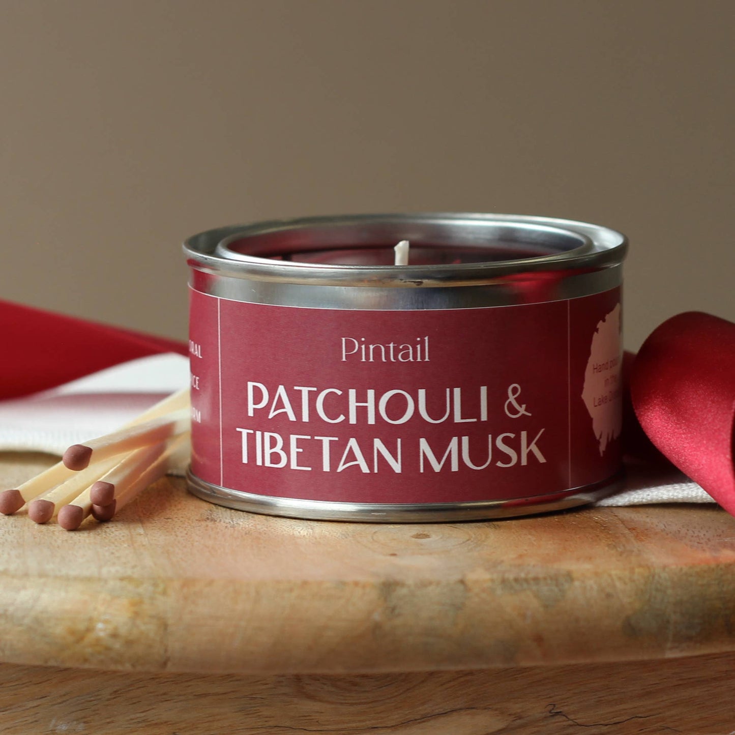 Candle in a metal tin labeled 'Patchouli & Tibetan Musk' by Pintail on a wooden surface with matches and red ribbons.