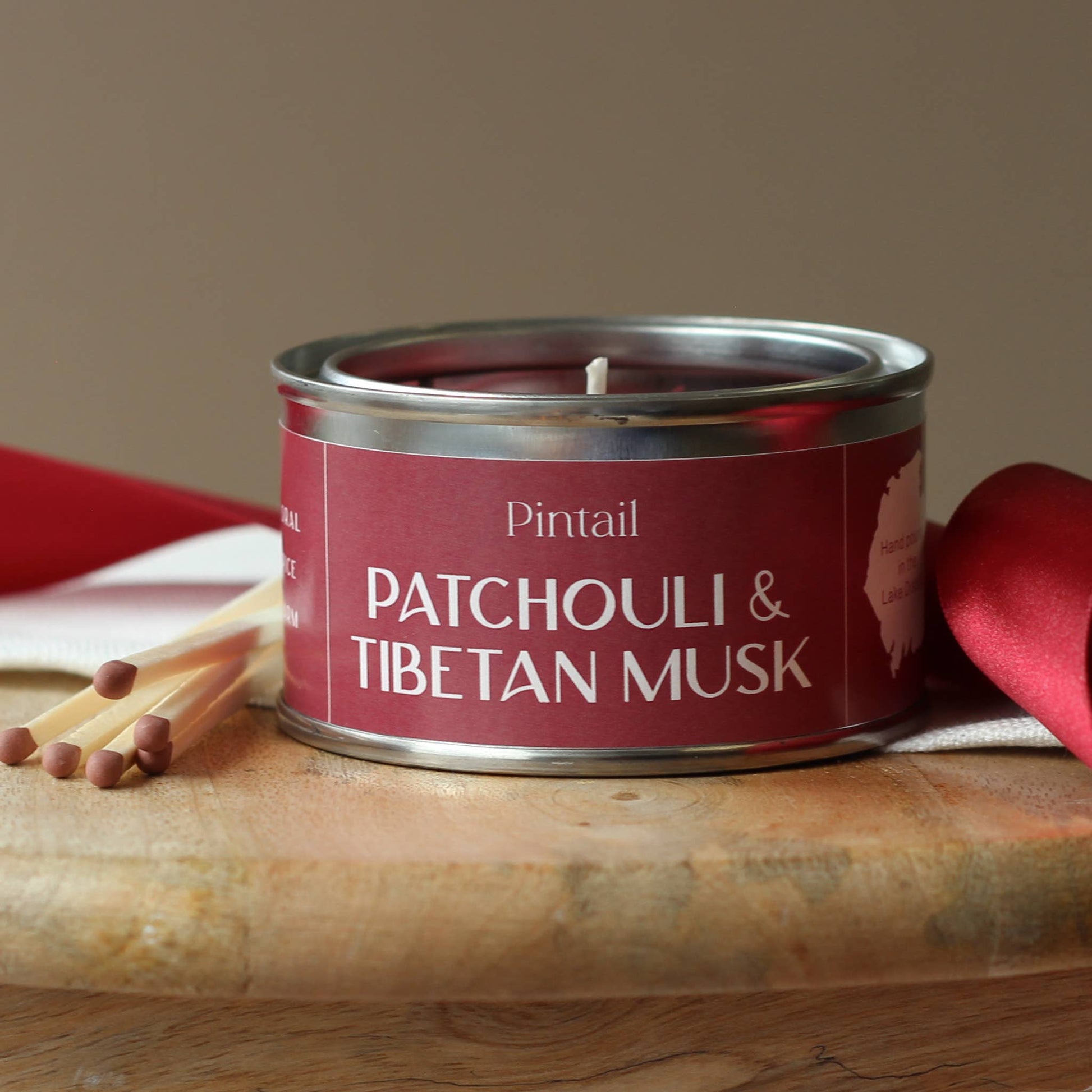 Candle in a metal tin labeled 'Patchouli & Tibetan Musk' by Pintail on a wooden surface with matches and red ribbons.