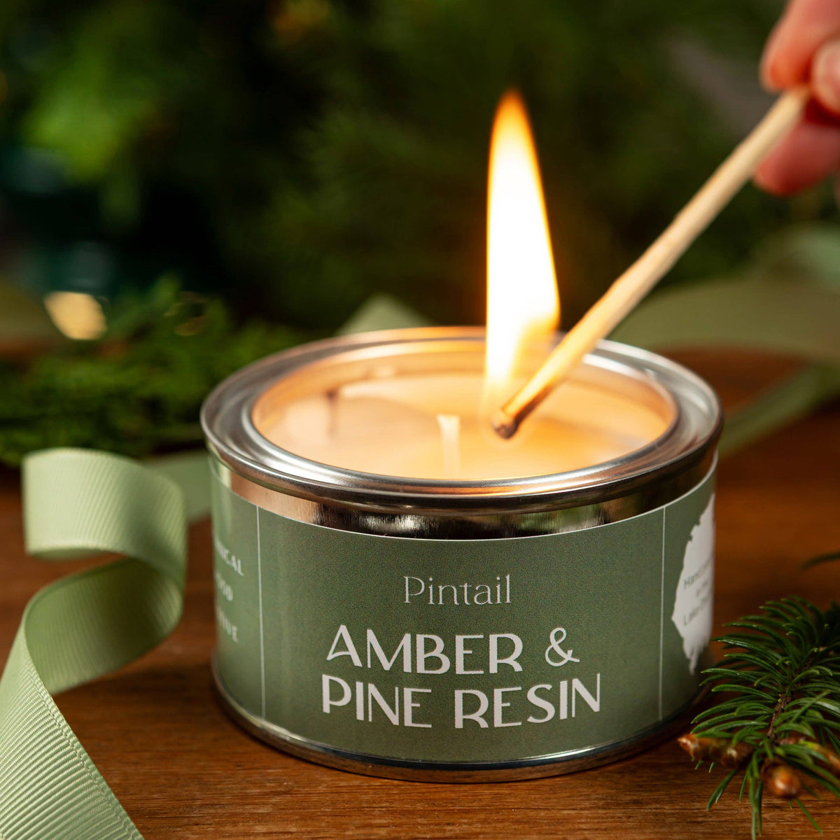 A lit candle in a round metal tin labeled 'Pintail AMBER & PINE RESIN' sits on a wooden surface with a match being held over the flame by a hand. A pale green ribbon and pine branches are visible in a softly blurred holiday setting.