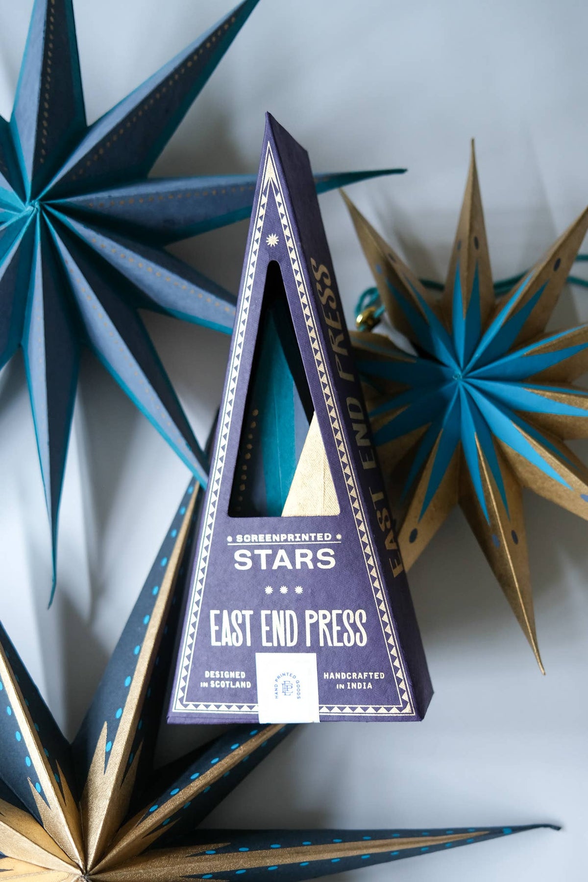 A triangular box for East End Press 'Stars' confectionery product, styled among large decorative paper star ornaments in blue, gold, and navy on a light gray background.