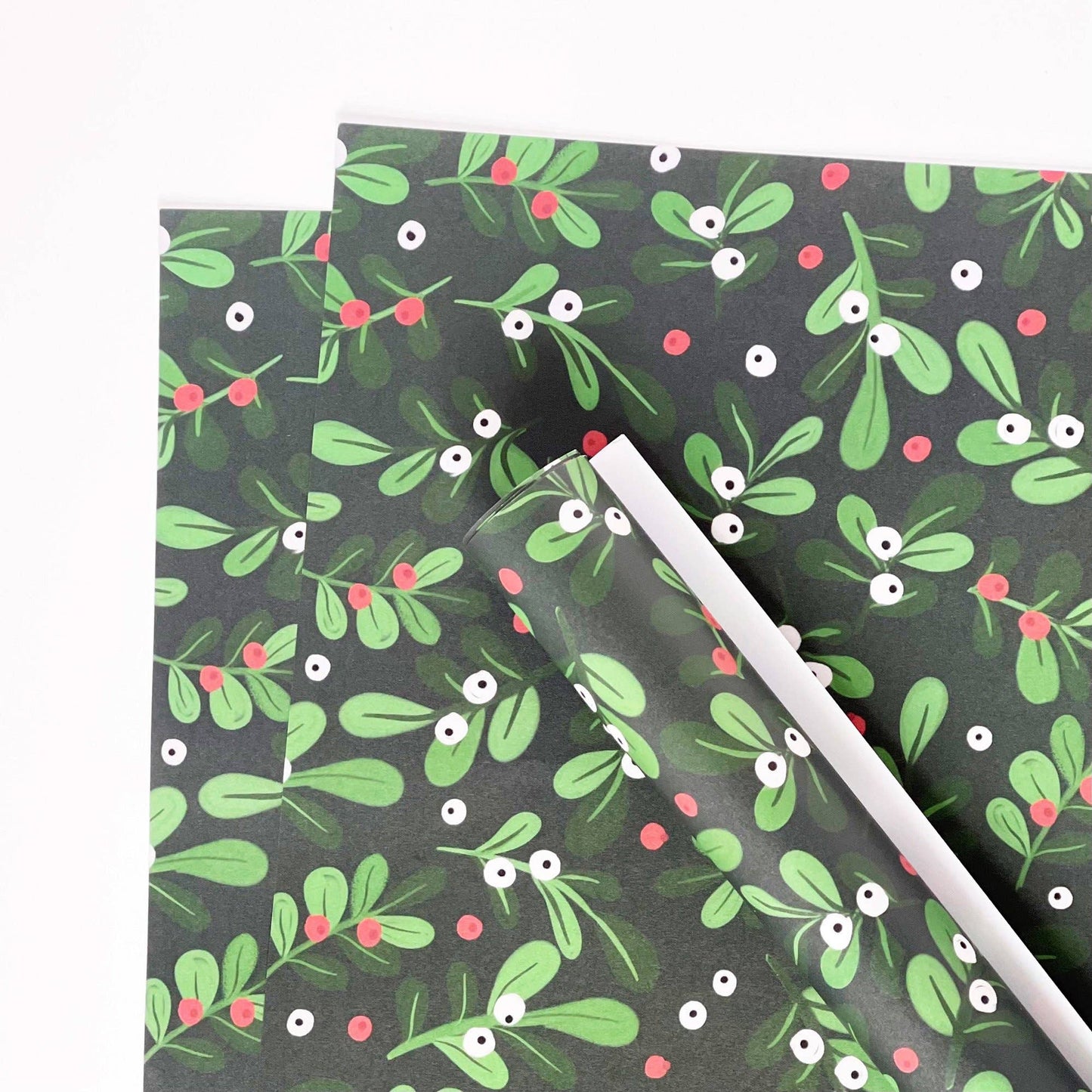 A sheet of wrapping paper with a dark gray background and a festive green mistletoe pattern, featuring red berries and white-and-black eyes. A second sheet is partially visible underneath, and a clear plastic roll is unwrapping the pattern.