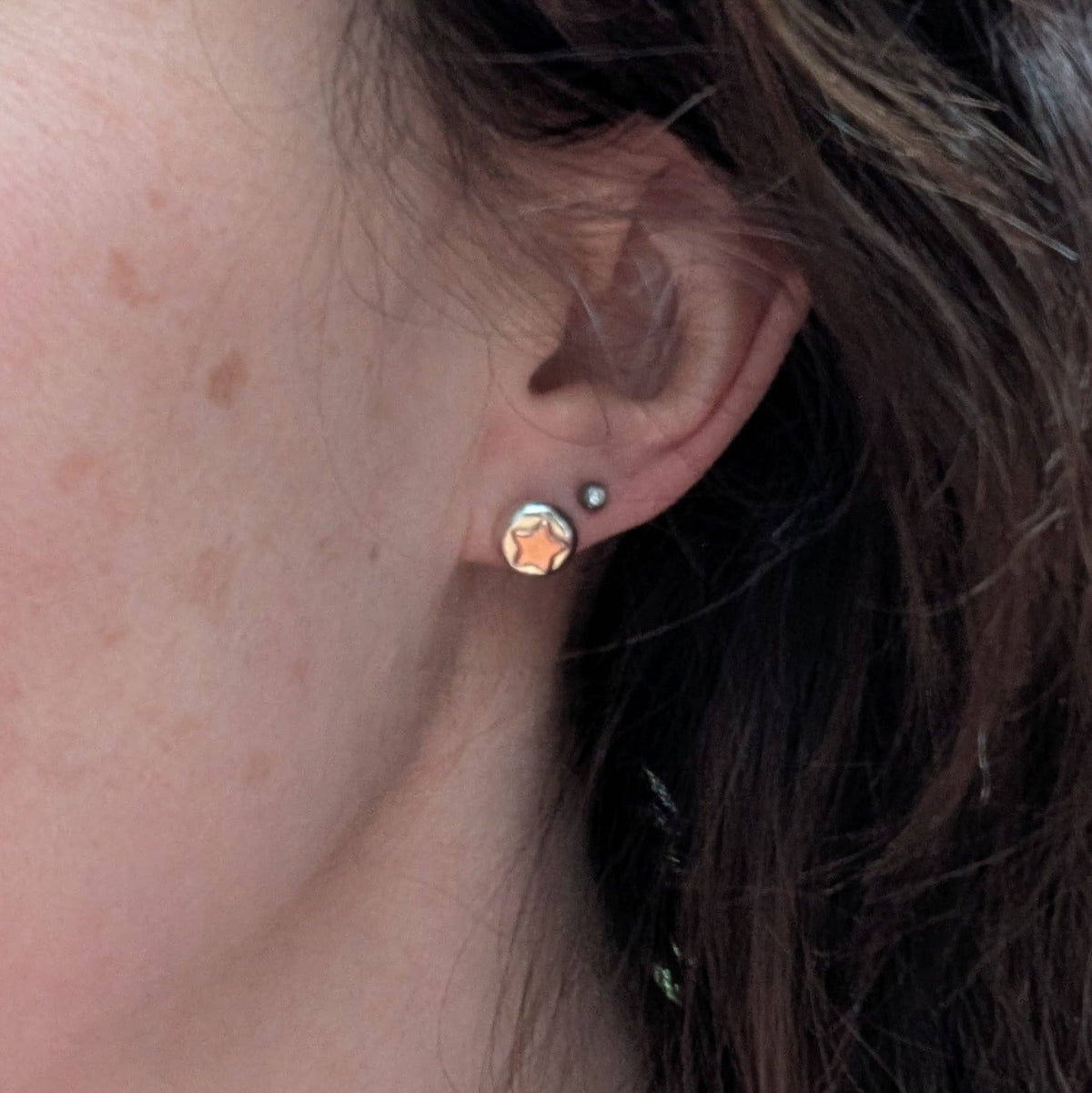 A close-up of a person's ear wearing a small silver stud earring with a faceted star-shaped crystal that reflects warm gold and peach tones, alongside a smaller silver hoop earring.
