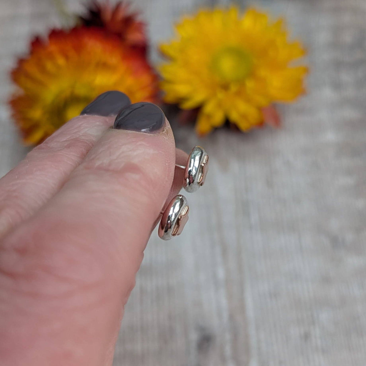 A close-up of a hand holding a pair of small silver hoop earrings against a blurred background with yellow and orange flowers.