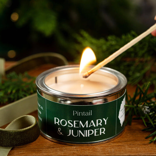 Candle labeled 'Rosemary & Juniper' being lit with a match, surrounded by greenery.