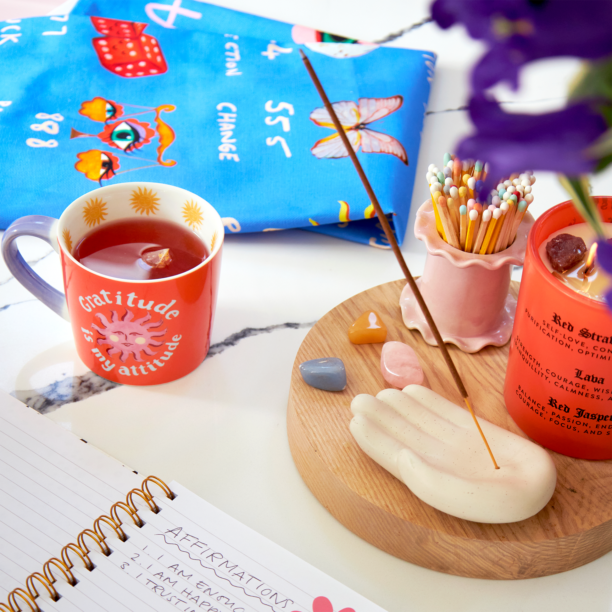 Red mug with 'Gratitude is my attitude' text, incense stick on a wooden coaster, and colorful pencils in the background.