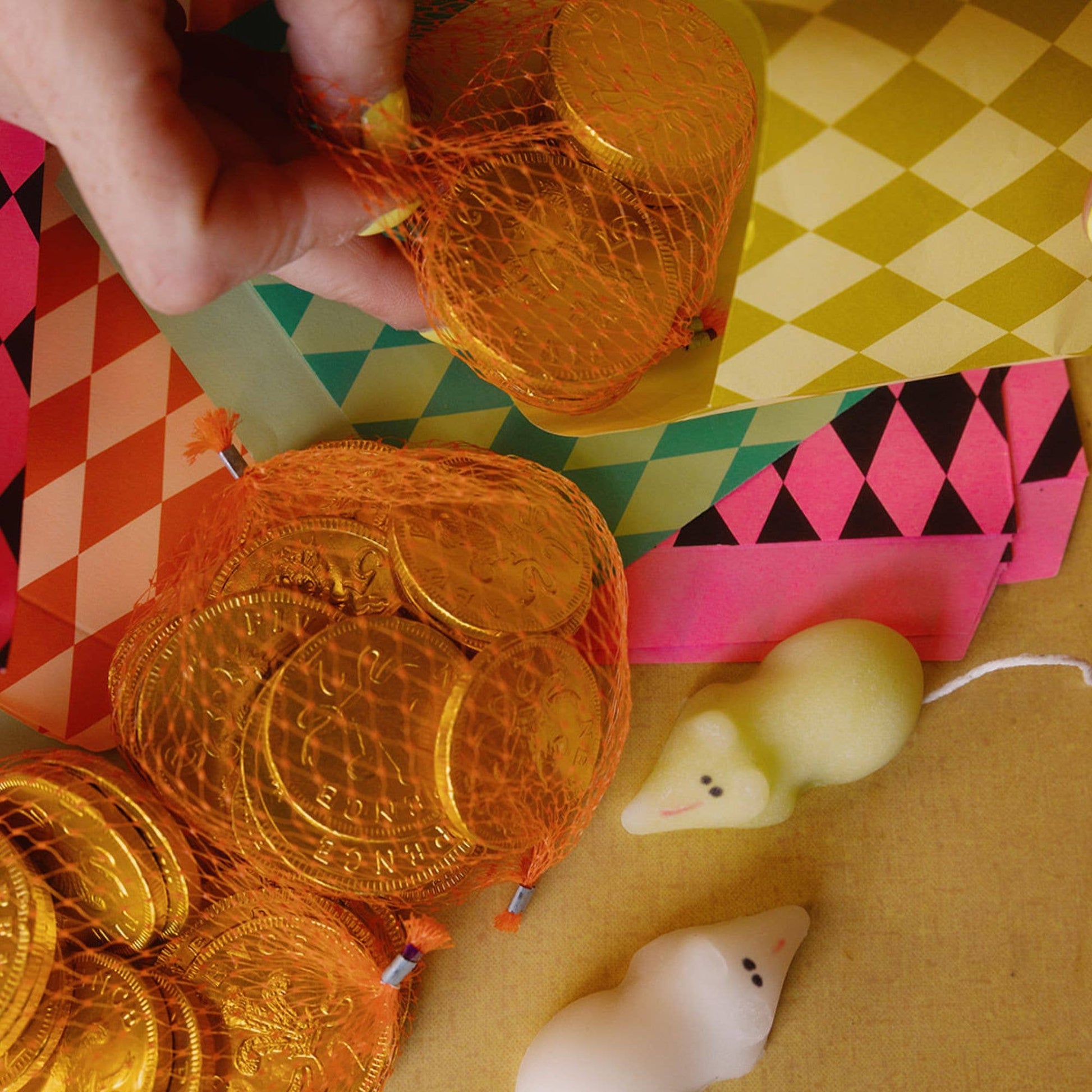 Hand holding gold coins in orange net bags on a colorful geometric-patterned surface.