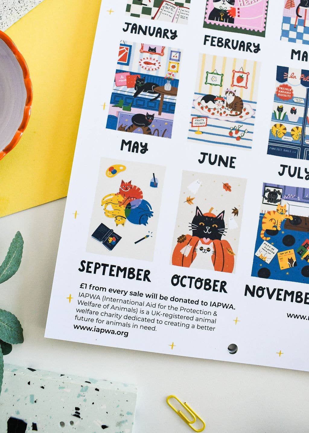 A printed monthly calendar page with colorful illustrated cat scenes and text stating donations to LAPWA, photographed on a white surface with a yellow envelope and small paperclip nearby.