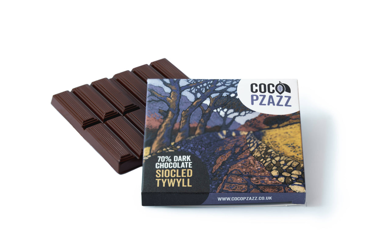 A bar of dark chocolate with its printed wrapper in front, photographed on a clean white background. The wrapper shows the brand name COCO PZAZZ and text reading 70% DARK CHOCOLATE SIOCLED TYWYLL, with a scenic landscape illustration.