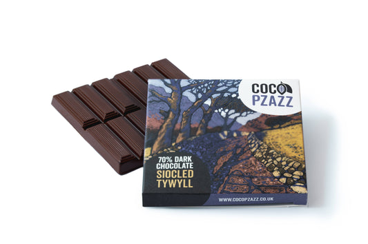 A bar of dark chocolate with its printed wrapper in front, photographed on a clean white background. The wrapper shows the brand name COCO PZAZZ and text reading 70% DARK CHOCOLATE SIOCLED TYWYLL, with a scenic landscape illustration.