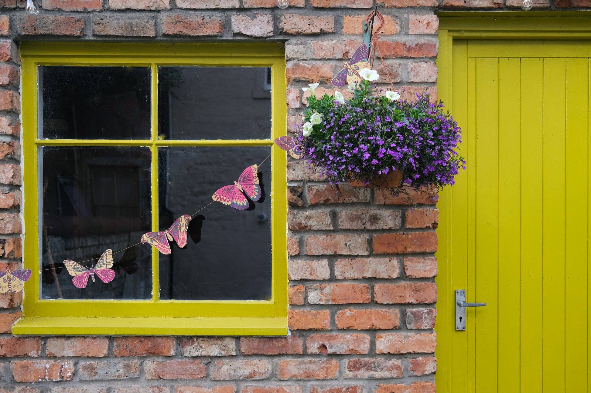 Brick wall with a yellow-framed window, pink butterfly decorations, and a hanging basket of flowers.