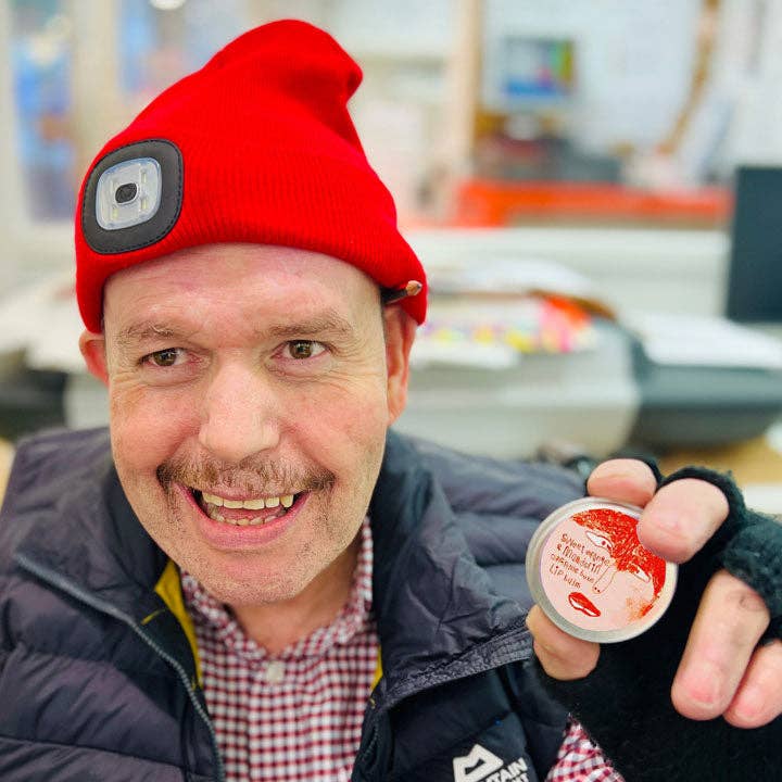 Man wearing a red beanie with a camera lens and holding a small container outdoors.