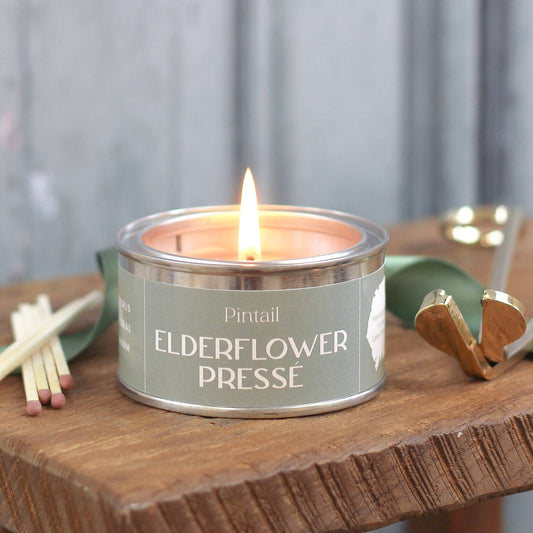 Candle in a tin labeled 'Elderflower Presse' by Pintail, lit on a wooden surface with a blurred background.