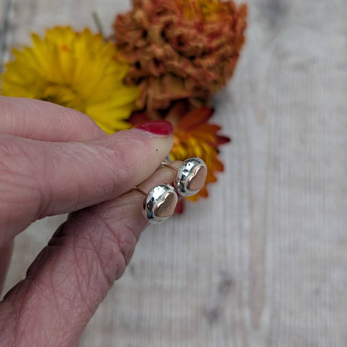 A close-up of a hand holding a pair of silver-toned stud earrings with oval clear stones, set against a rustic light wood background with yellow and orange flowers.