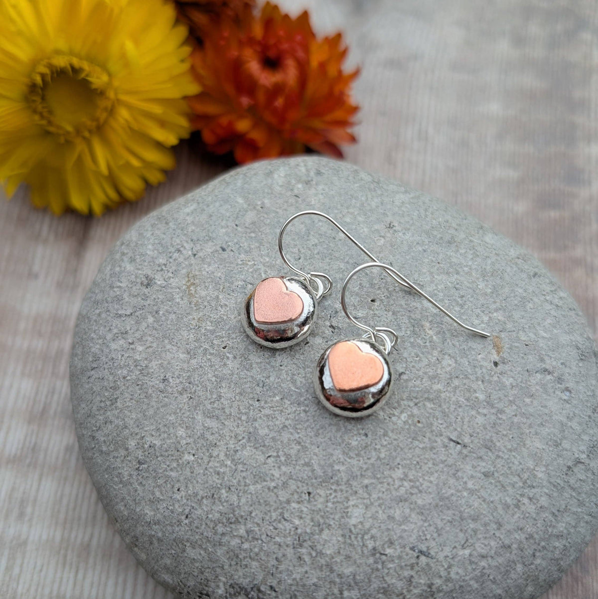 A pair of silver dangle earrings with small oval charms featuring a pink heart in the center, displayed on a round gray stone with yellow and orange flowers in the background.
