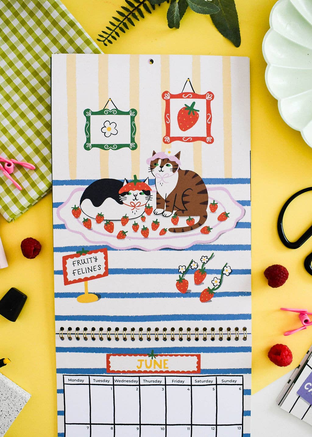A decorative hanging calendar page with illustrated cats, strawberries, and a June monthly grid, styled on a bright yellow surface with small props around it.