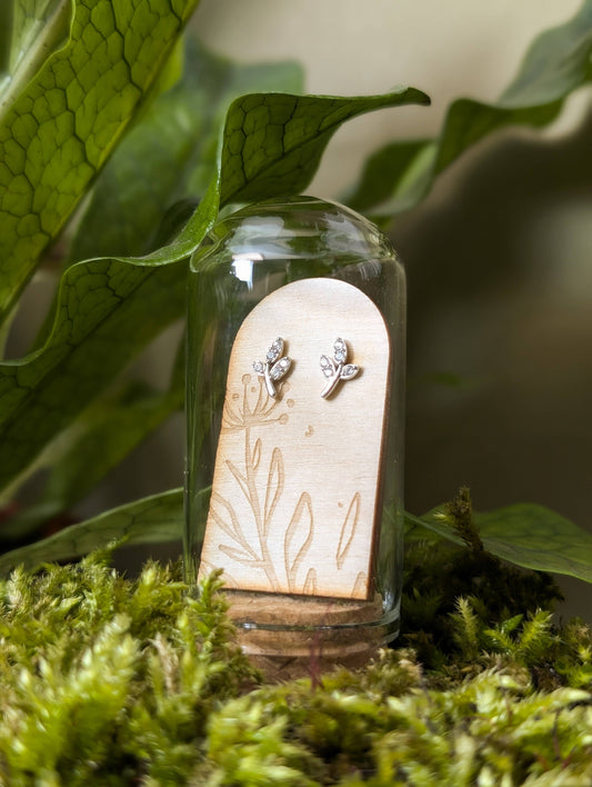 Sterling silver and cubic zirconia leaf-shaped stud earrings displayed inside a glass terrarium tube, placed on a bed of moss with green leaves in the background.