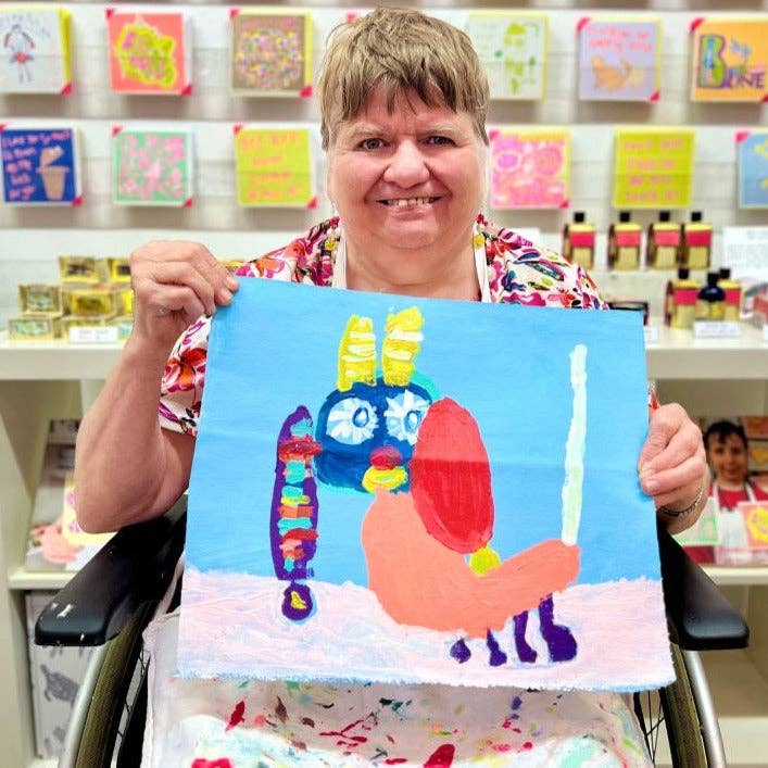 Person in a colorful shirt holding a child's drawing of a colorful creature in a store setting.