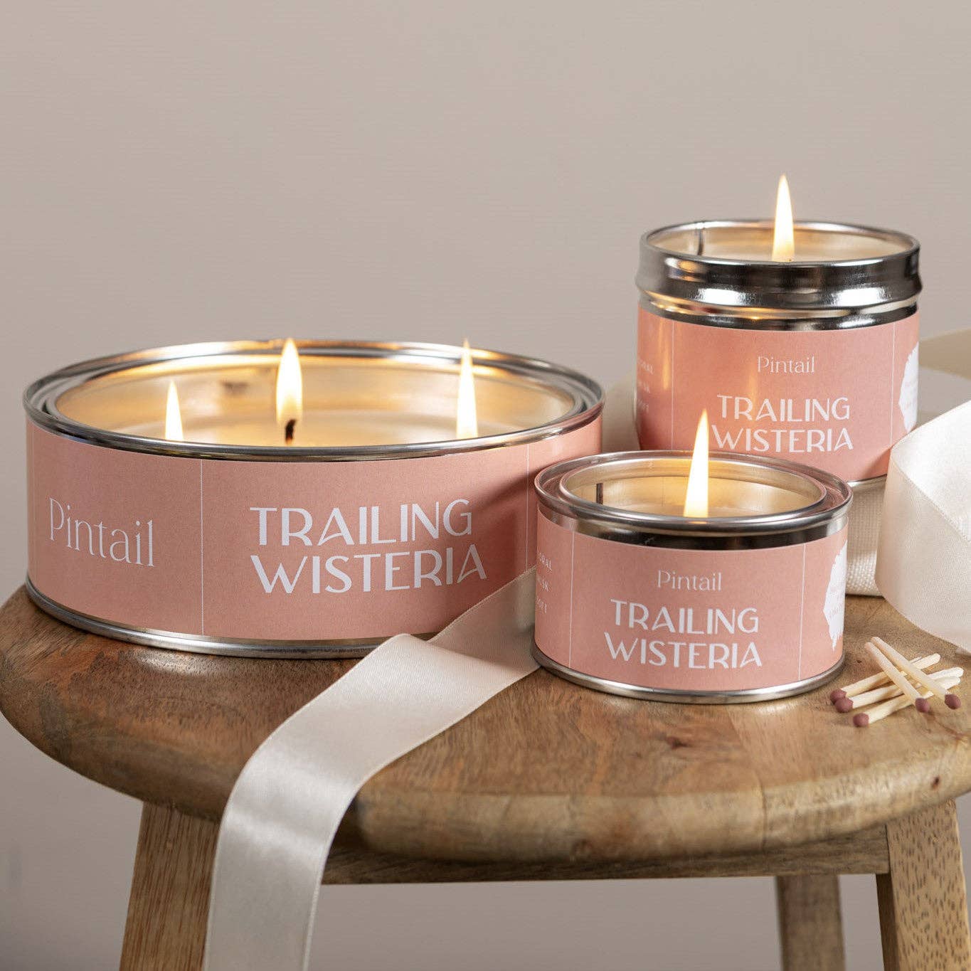 Candle in a tin labeled 'Trailing Wisteria' by Pintail on a wooden surface with white fabric.