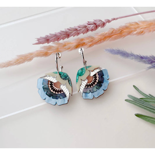 A pair of bohemian-style flower dangle earrings with layered blue, green, and white enamel petals and silver hooks, displayed on a white surface with dried decorative grasses.