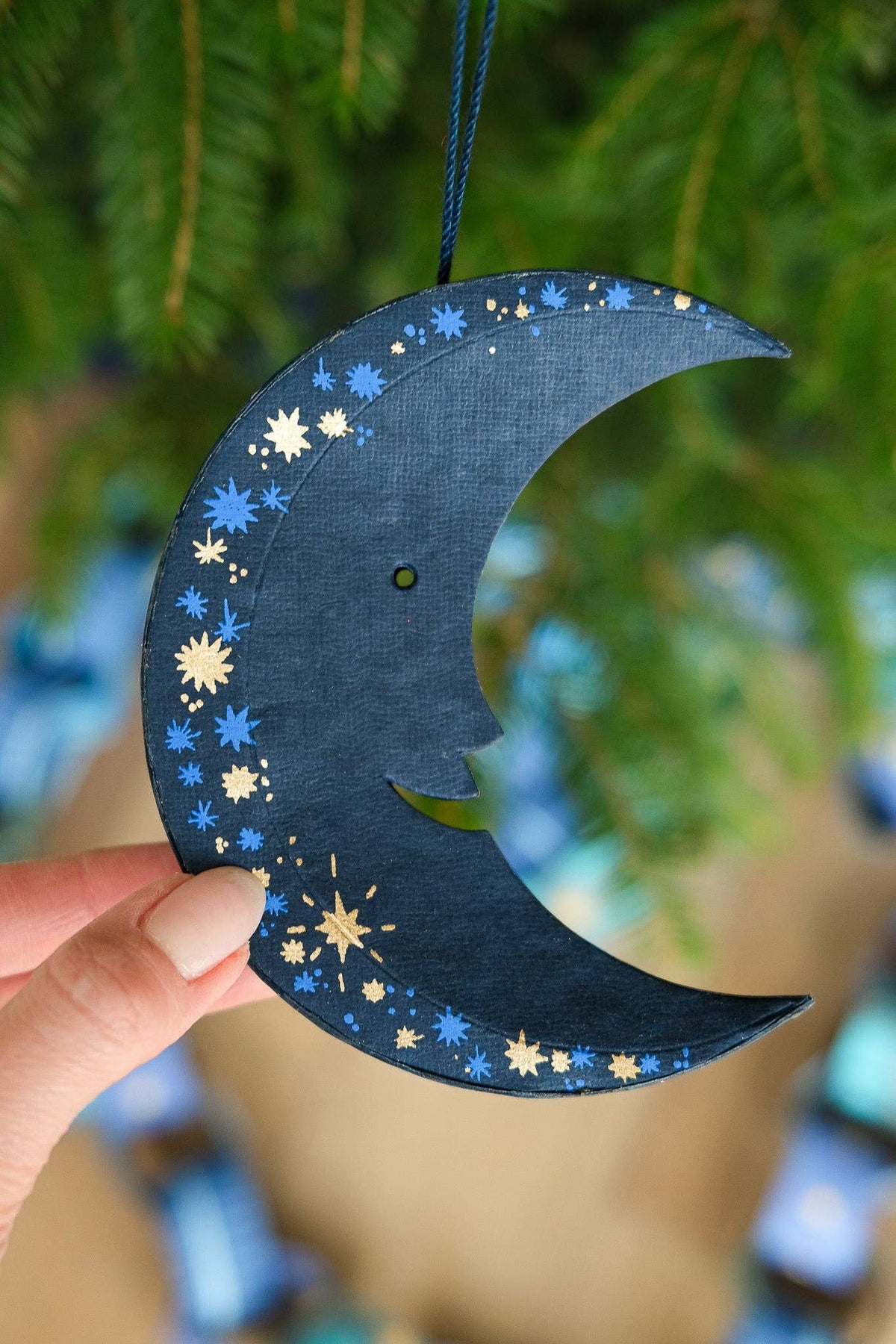 Decorative blue crescent moon ornament with star patterns held against a blurred green background.