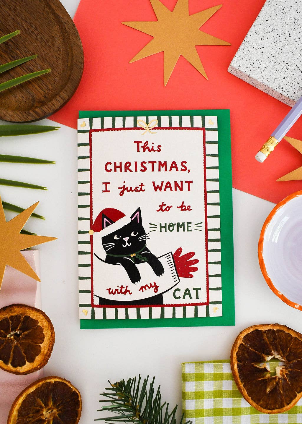 A Christmas greeting card featuring a black cat wearing a red Santa hat and holding a red paw print, with the text 'This Christmas, I just WANT to be HOME with my CAT'. The card is styled in a flat-lay arrangement with dried orange slices, pine needles, gold star cutouts, and other holiday props on a white surface.