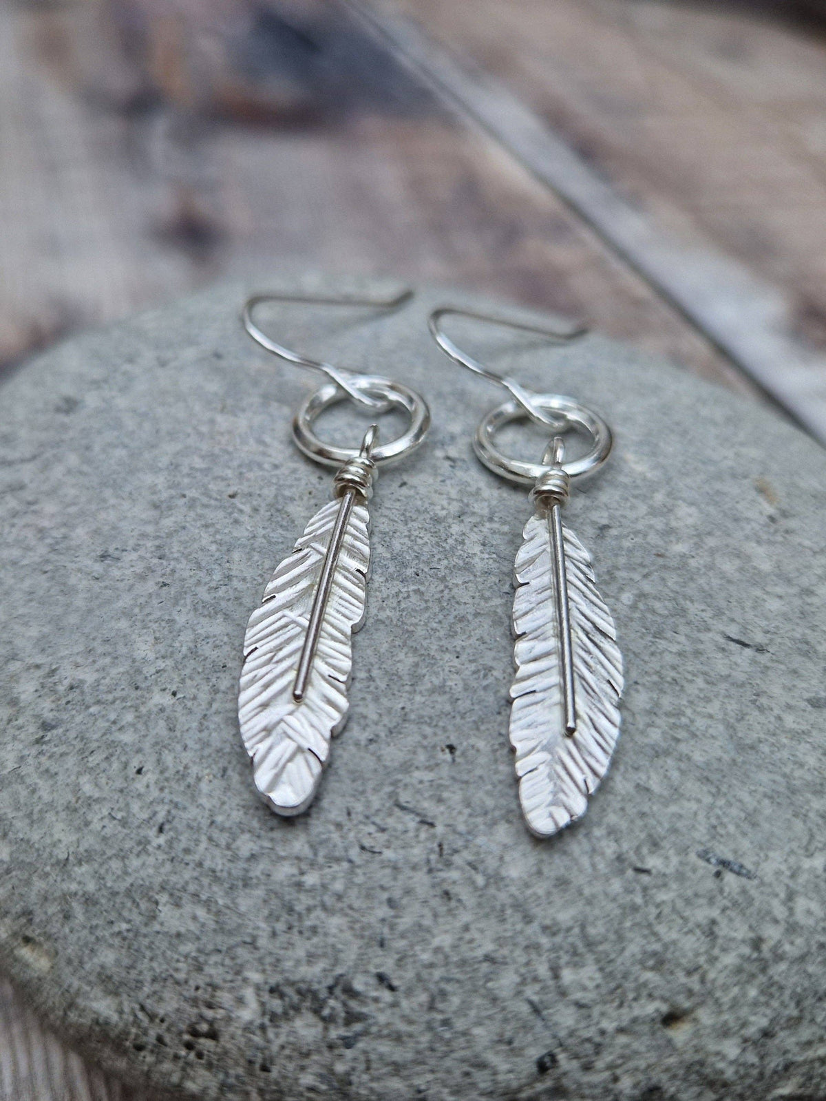 A pair of silver feather drop earrings with textured, hammered details, displayed on a smooth gray stone over a rustic wooden surface.
