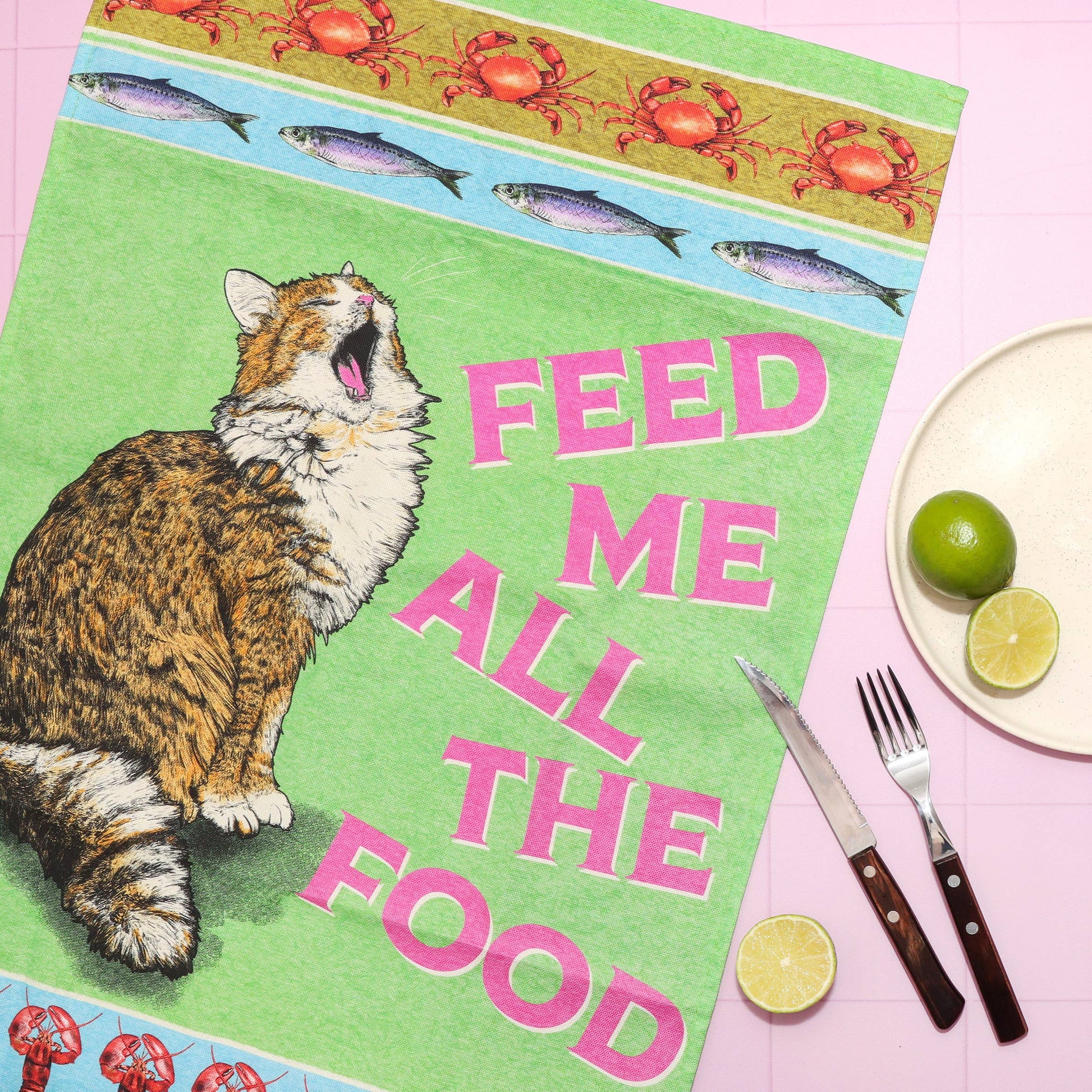 Cat-themed placemat with text 'Feed Me All The Food' on a pink background with cutlery and limes.