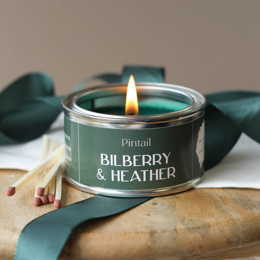 Candle in a tin labeled 'Bilberry & Heather' with matches and ribbons on a wooden surface.