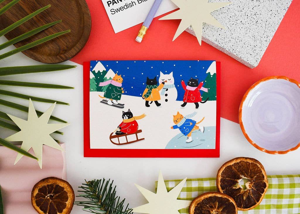 A square Christmas greeting card featuring illustrated cats playing outdoors in winter clothing, surrounded by flat-lay props including dried orange slices, a small bowl, a notebook, and decorative stars on a red and white surface.