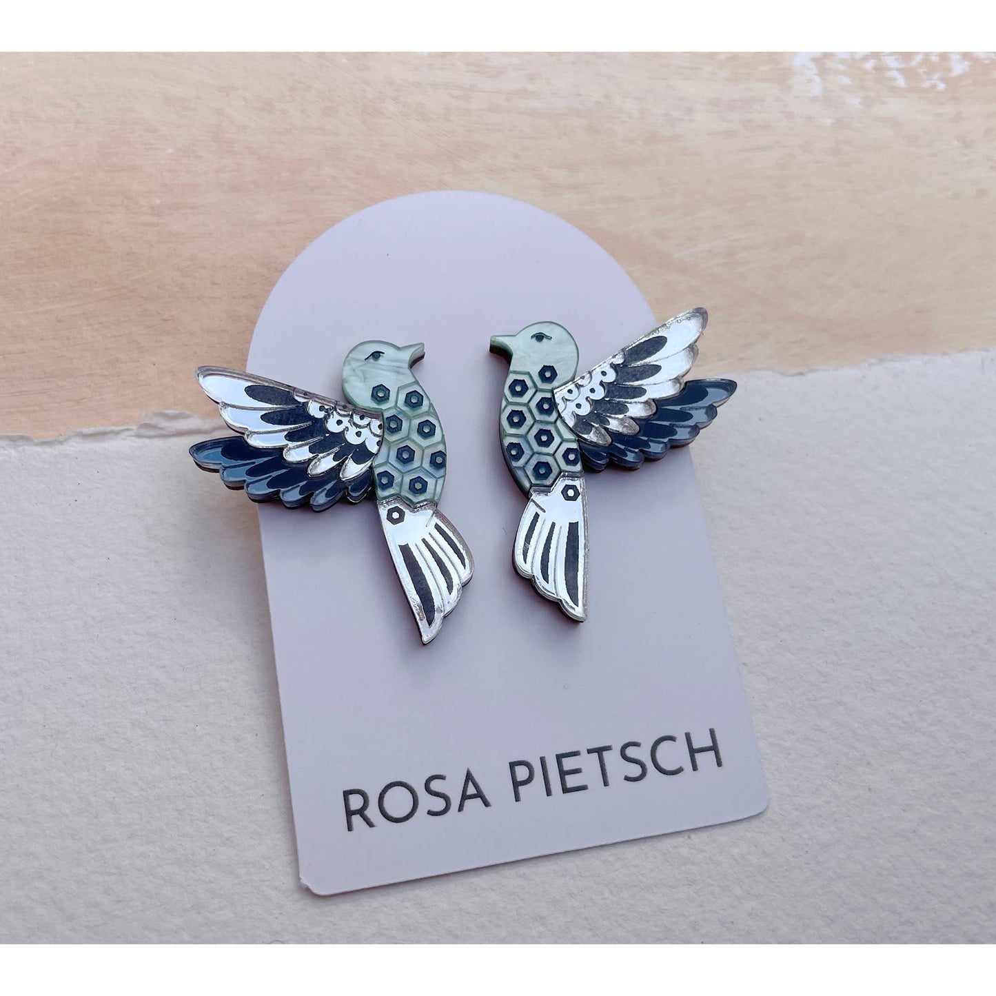A pair of bird-shaped earrings displayed on a lavender card labeled ROSA PIETSCH. The earrings have a silver-tone metal base with black, white, and light blue enamel details, set against a split beige and off-white textured background.