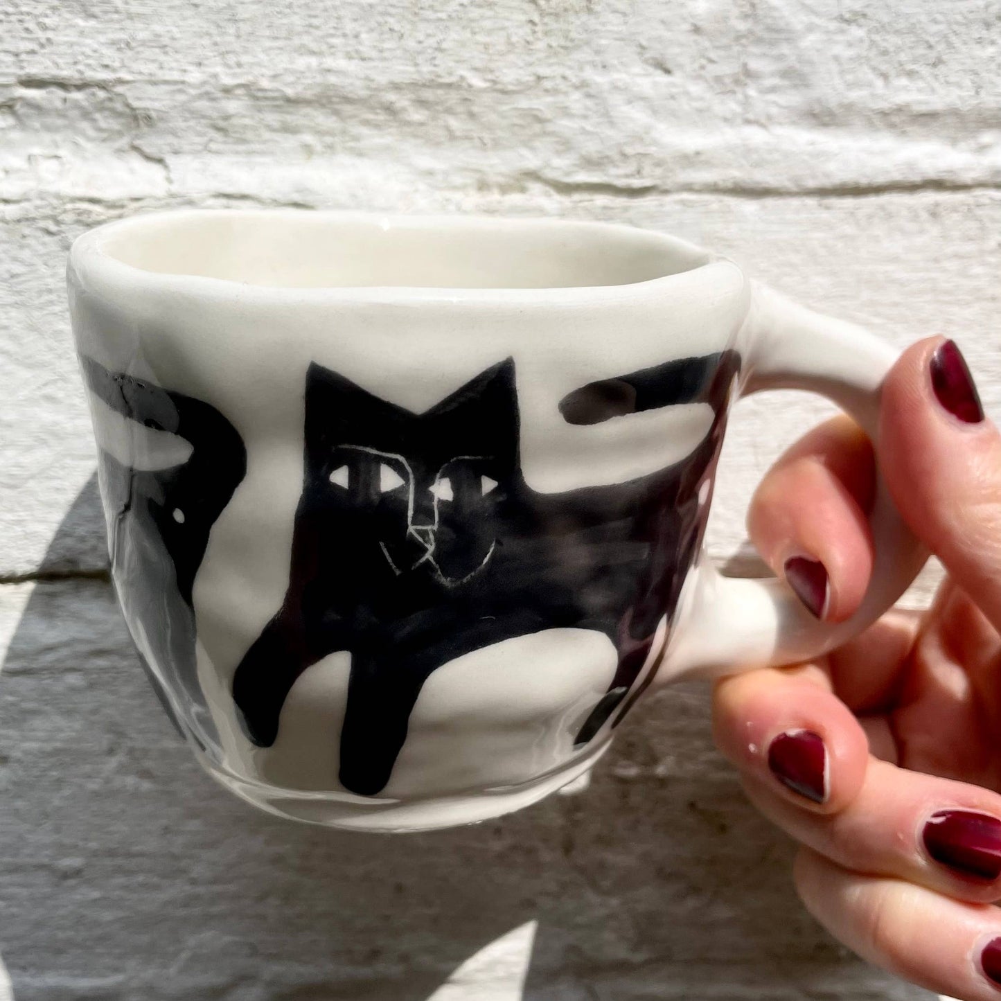 White mug with black cat design held by a hand with red nail polish on a light wooden surface.