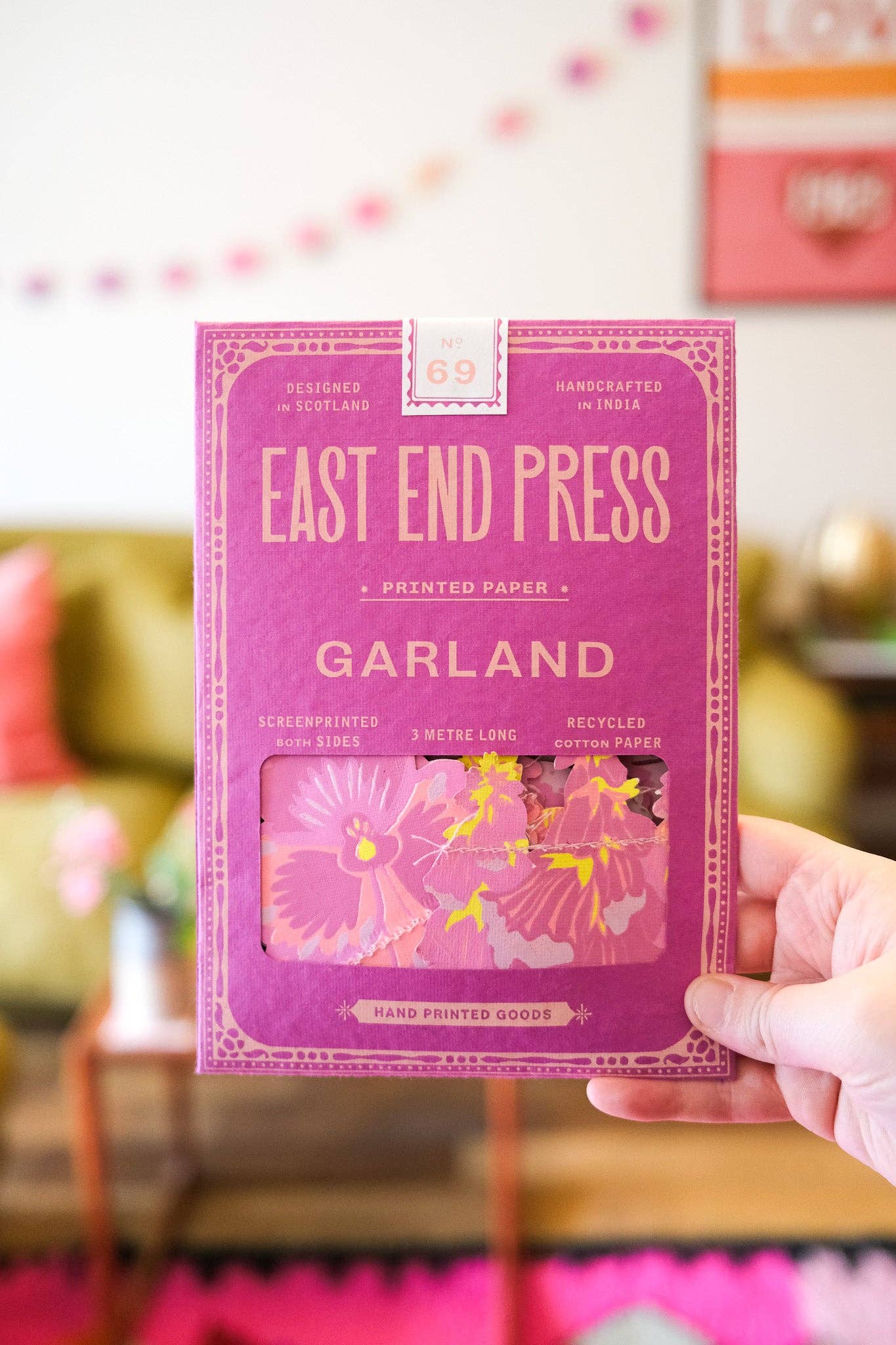 Hand holding a pink package labeled 'East End Press Garland' with a blurred indoor background.