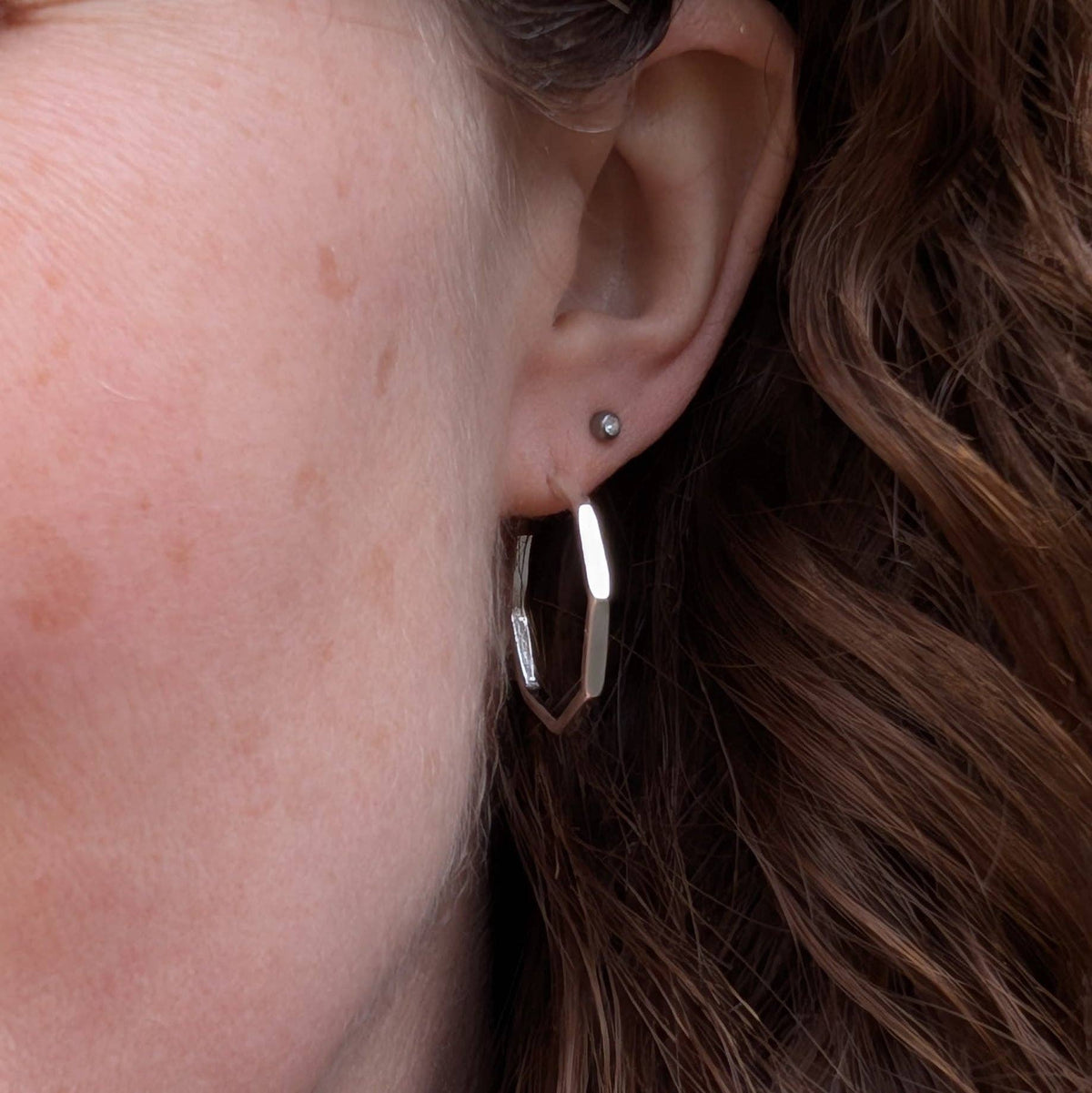 A close-up of a person's ear wearing a polished silver geometric hoop earring and a small silver stud earring, with brown hair visible.