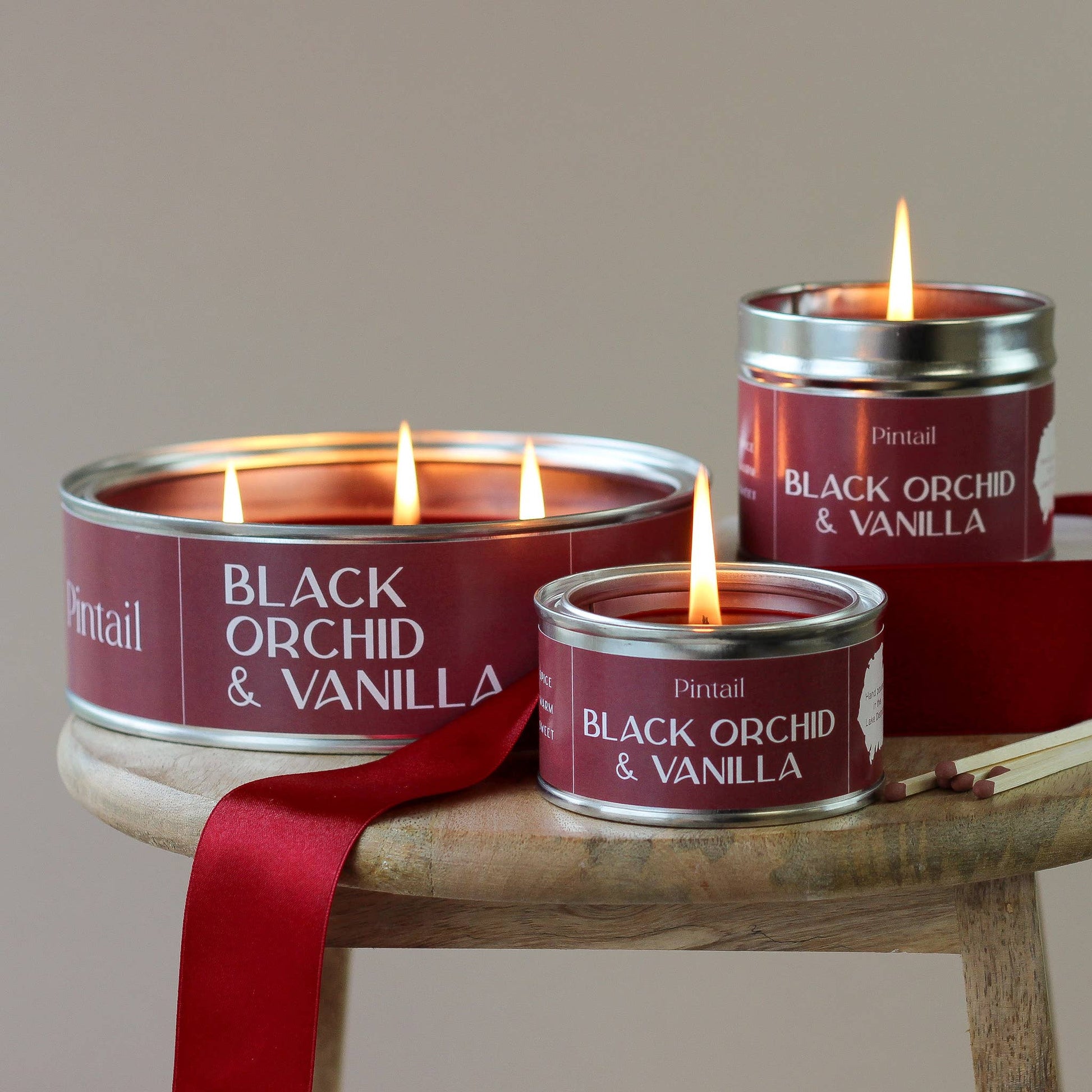 Three candles labeled 'Black Orchid & Vanilla' on a wooden stand with a red ribbon.