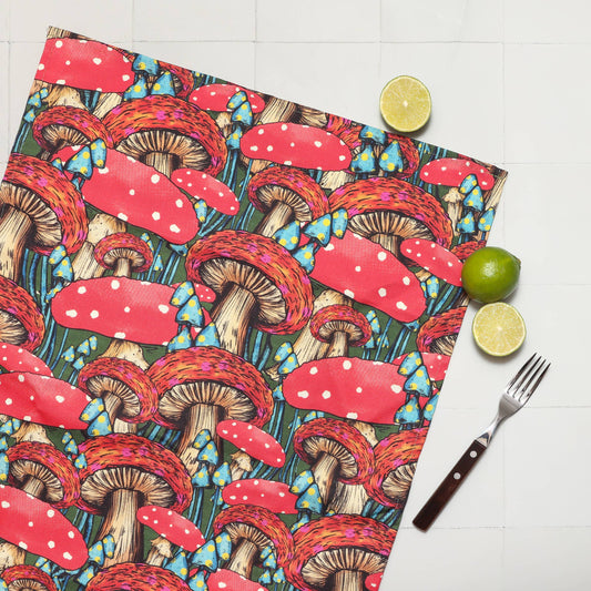 A square fabric item, likely a napkin or small tablecloth, featuring a colorful mushroom print, styled flat-lay on a white tiled surface with a fork, lime, and a slice of lime.