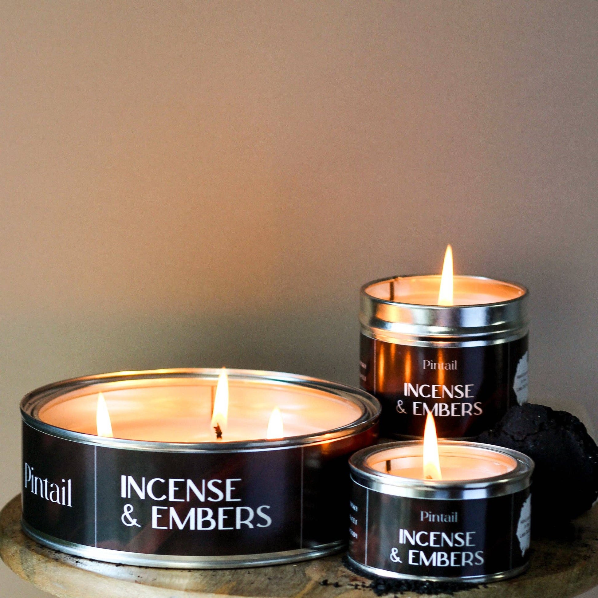 Three 'Incense & Embers' candles in tins on a wooden surface with a neutral background.