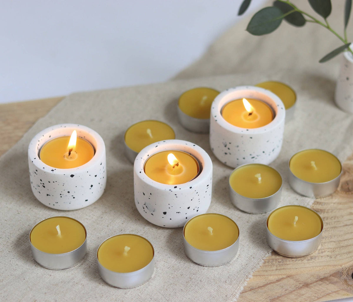 Yellow candles in white holders on a wooden surface with a plant in the background