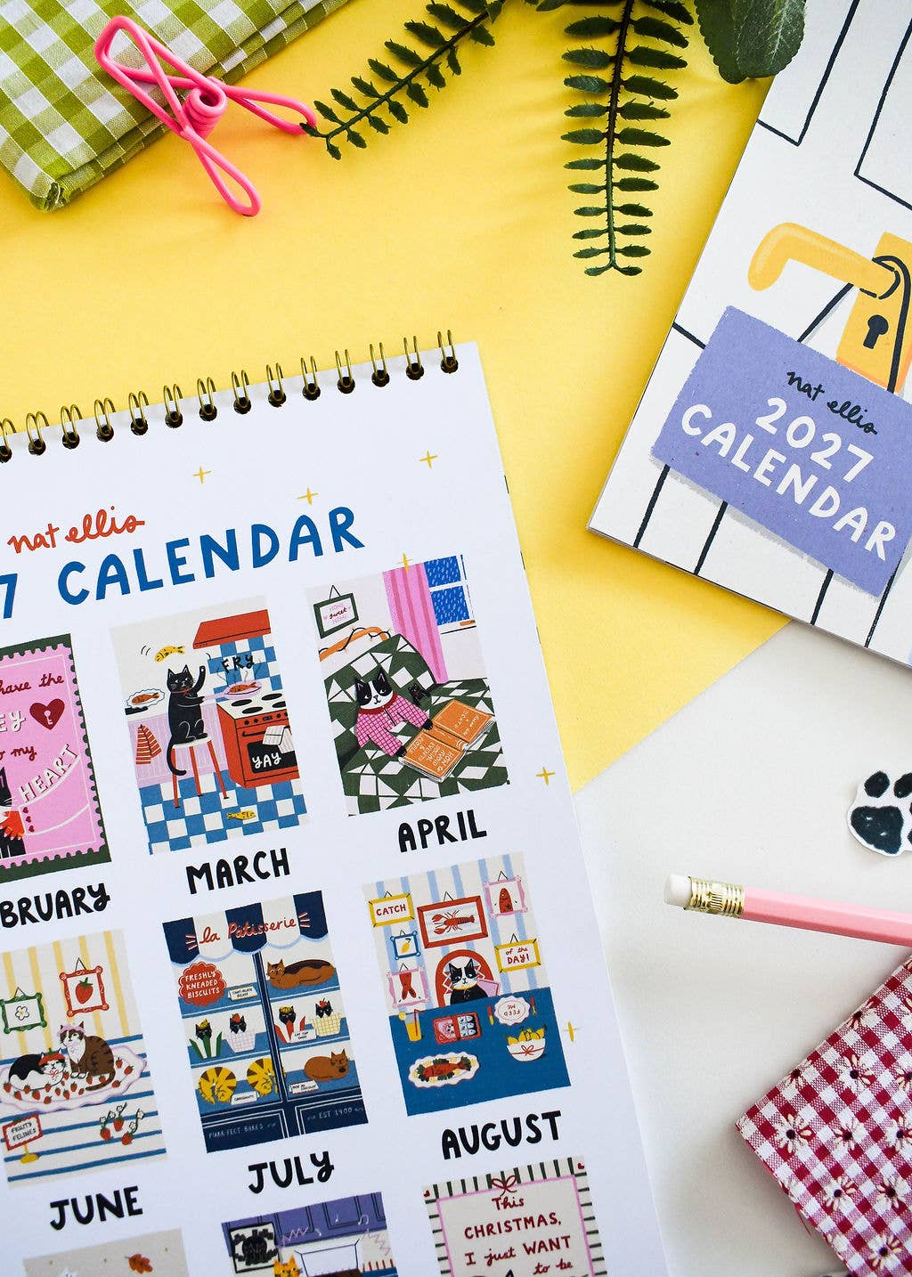 A spiral-bound 2027 calendar page from not ella is shown in a styled flat-lay with other stationery items including a pink pencil, notebook, and ribbon on a yellow surface.