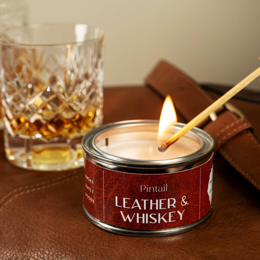 Candle labeled 'Leather & Whiskey' being lit with a glass of whiskey in the background.