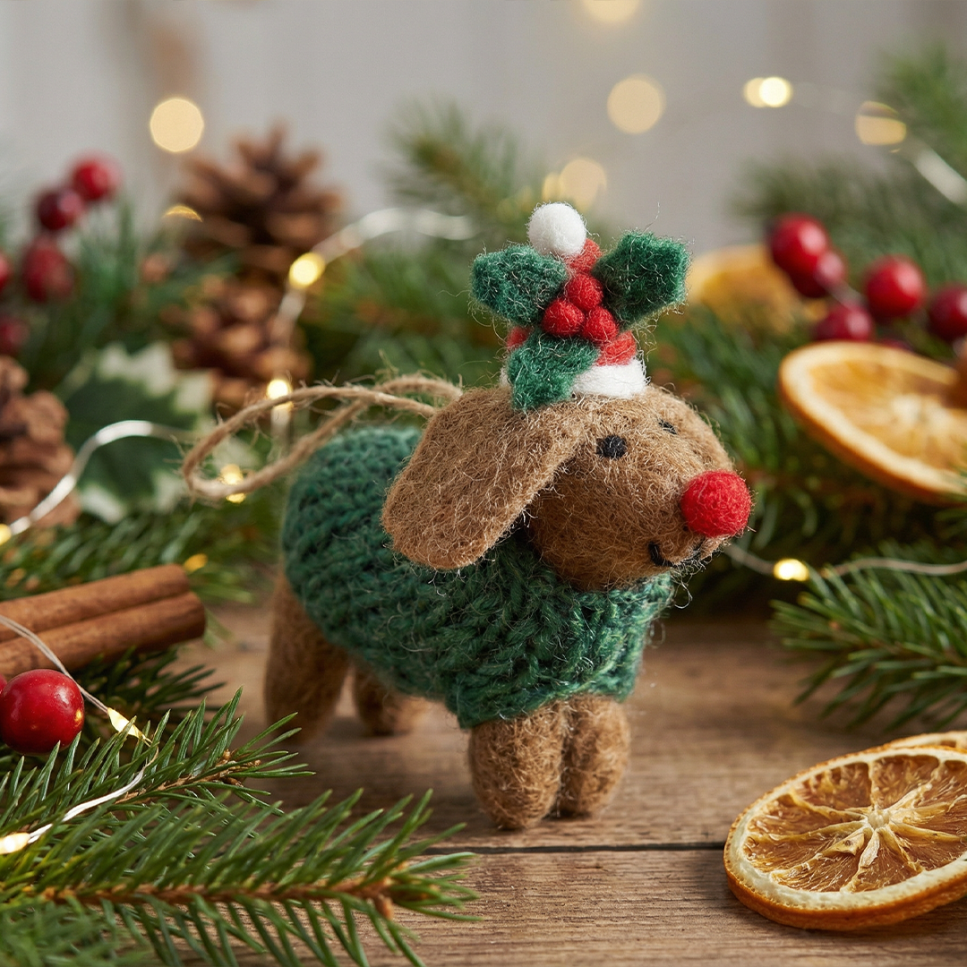 A handmade felt Christmas ornament shaped like a brown dog wearing a green knitted sweater and a festive hat with red berries, displayed on a wooden surface surrounded by pine branches, dried orange slices, cinnamon sticks, and warm fairy lights.