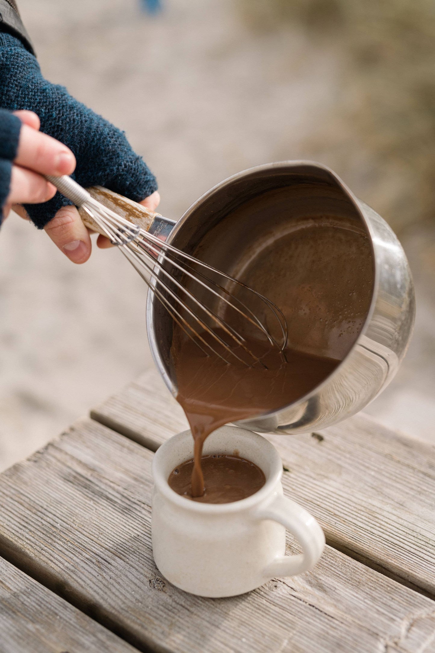 Person pouring chocolate mixture from a metal pot into a white mug on a wooden surface.