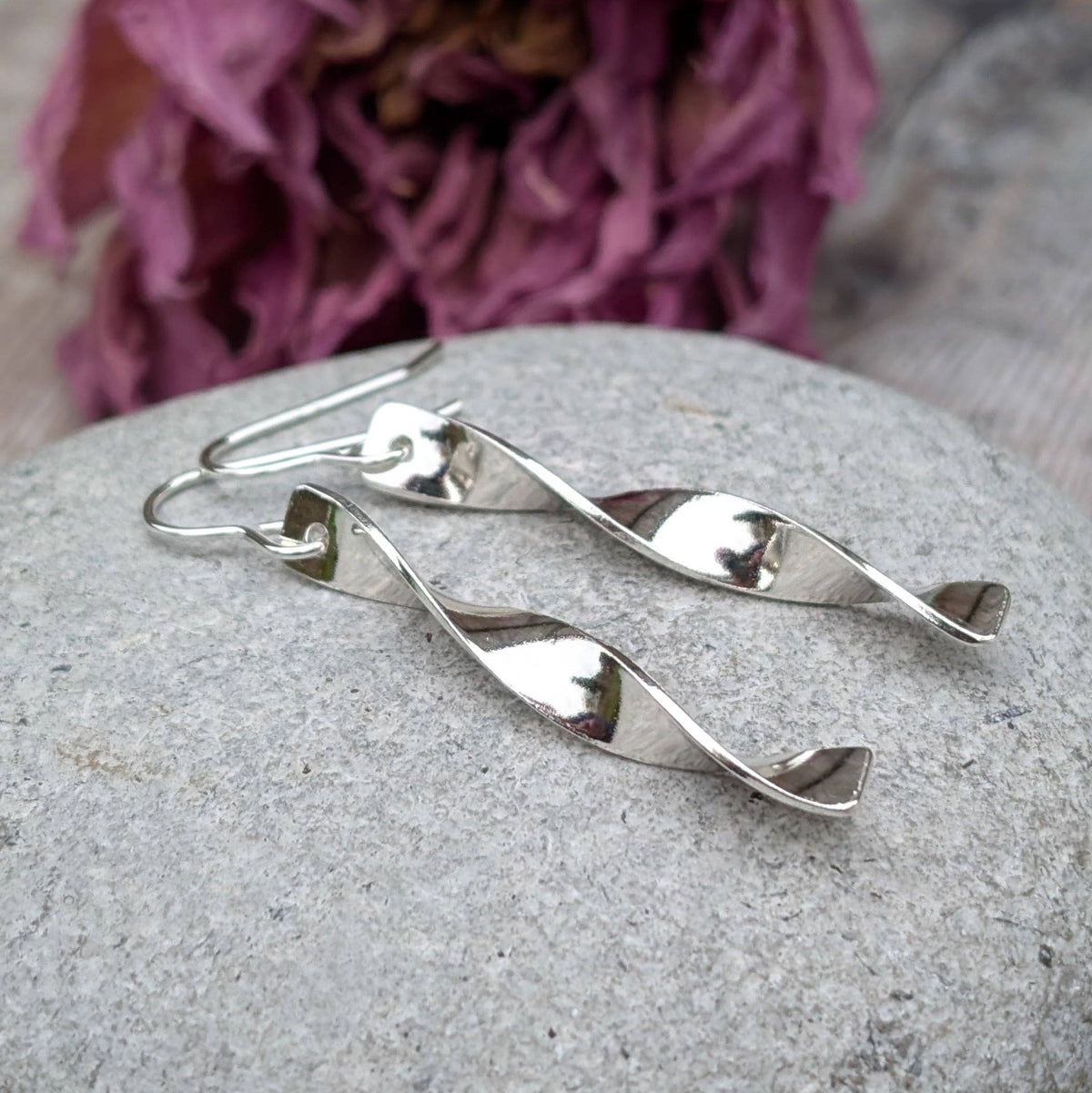A pair of polished silver dangle earrings with a twisted spiral design, resting on a textured gray stone with a blurred purple flower in the background.