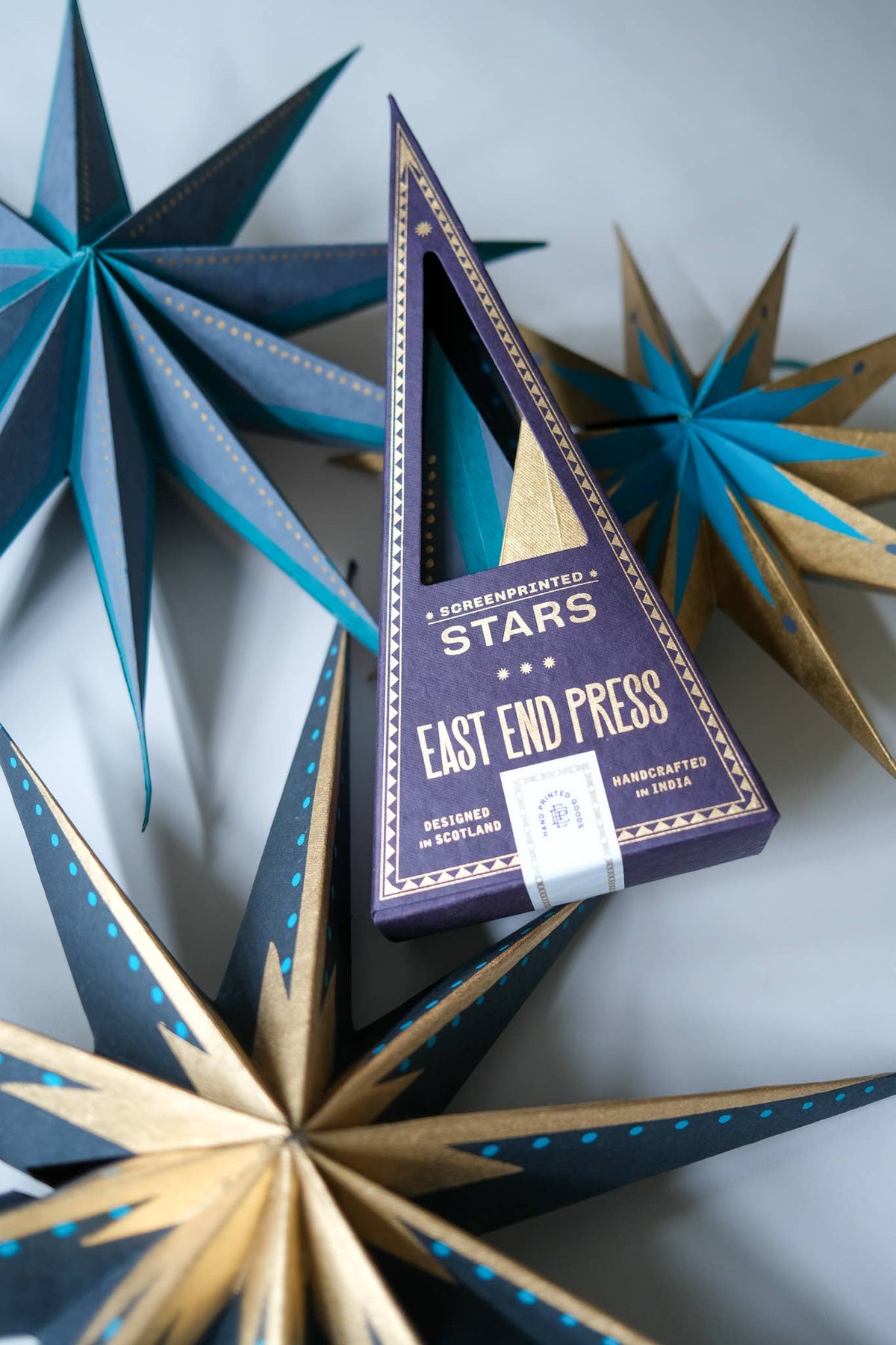 A triangular purple box labeled 'EAST END PRESS' and 'SPECKLED STARS' is photographed on a light gray surface surrounded by decorative star-shaped ornaments in navy, teal, and gold.